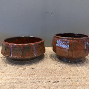 May include: Two handmade ceramic bowls with a rich, earthy color palette. The bowls feature a reddish-brown glaze with darker accents, and a textured, faceted design. The bowls are displayed on a natural fiber surface.