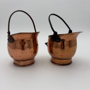 May include: Two small copper coal scuttles with rounded bodies, flared rims, and dark metal swing handles. Each vessel features a polished metallic finish with visible surface texture and a sturdy base, designed with a wide pouring spout.