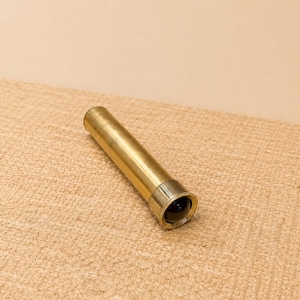 May include: A brass telescope with a dark lens, lying on a tan woven surface. The telescope is cylindrical with a slightly wider end. The brass has a polished, reflective finish.