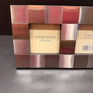 May include: A rectangular multi-opening picture frame constructed from brushed metal squares in alternating silver and bronze tones. The frame features cream-colored inserts with the text michael graves at home and a barcode with 3x3 printed.