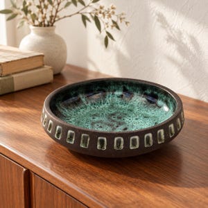 May include: A round ceramic bowl with a dark brown exterior featuring a repeating pattern of rectangular indentations. The interior of the bowl has a mottled glaze in shades of deep teal, turquoise, and black, creating a textured appearance.