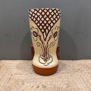 May include: A tall, ceramic vase with a beige background and brown floral design. The vase has a brown base and a lattice pattern at the top. Two small, textured handles are on the sides. The vase is approximately 10 inches tall.