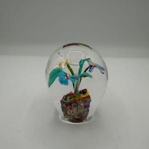 May include: A clear glass paperweight featuring an internal floral design with blue, green, and white petals. The base of the flower arrangement is composed of a textured, multi-colored speckled glass mound in shades of red, yellow, and blue.