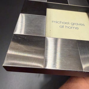 May include: A square picture frame featuring a checkerboard pattern of alternating brushed silver-tone metal and dark black squares. The center of the frame contains a cream-colored insert with the text michael graves at home printed in blue.