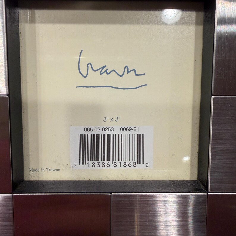 May include: A square picture frame with a brushed metallic finish. The cream-colored insert features a handwritten signature, the text 3" x 3", a barcode, the numbers 065 02 0253 0069-21, and the text Made in Taiwan printed in the bottom left corner.