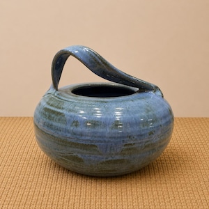 May include: A blue ceramic vase with a curved handle. The vase has a round body and a wide opening. The handle arches over the top. The glaze has a mottled appearance with shades of blue and green.