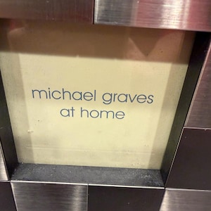 May include: A square sign with a light cream center featuring the blue text michael graves at home. The sign is framed by a border of alternating brushed silver and dark metallic square tiles arranged in a grid pattern around the central area.