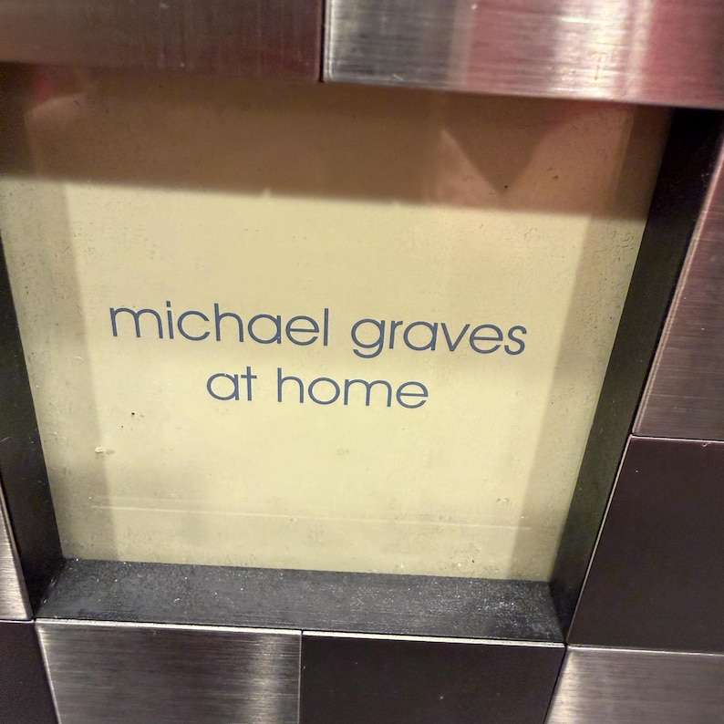 May include: A square sign with a light cream center featuring the blue text michael graves at home. The sign is framed by a border of alternating brushed silver and dark metallic square tiles arranged in a grid pattern around the central area.
