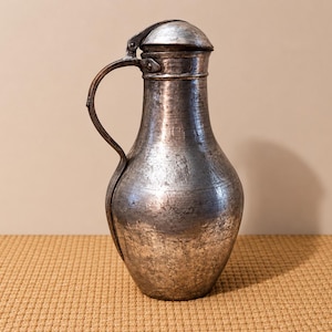 May include: A vintage metal pitcher with a hinged lid and curved handle. The pitcher has a tarnished, textured surface with a dark, aged appearance. The pitcher is likely made of copper or a similar metal.