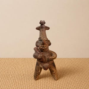 Pode incluir: Uma figura de cerâmica castanha e desgastada que retrata uma figura humana estilizada com uma figura menor no topo da cabeça. A figura está segurando um objeto menor. A figura tem uma textura áspera e fica em três pernas.