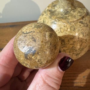 K&ouml;nnte beinhalten: Nahaufnahme einer runden, gesprenkelten Steinkugel, die in einer Hand gehalten wird, mit einer passenden dekorativen Vase im Hintergrund. Die Kugel und die Vase haben ein beige-braunes Farbschema mit poliertem Finish. Der Hintergrund ist unscharf.