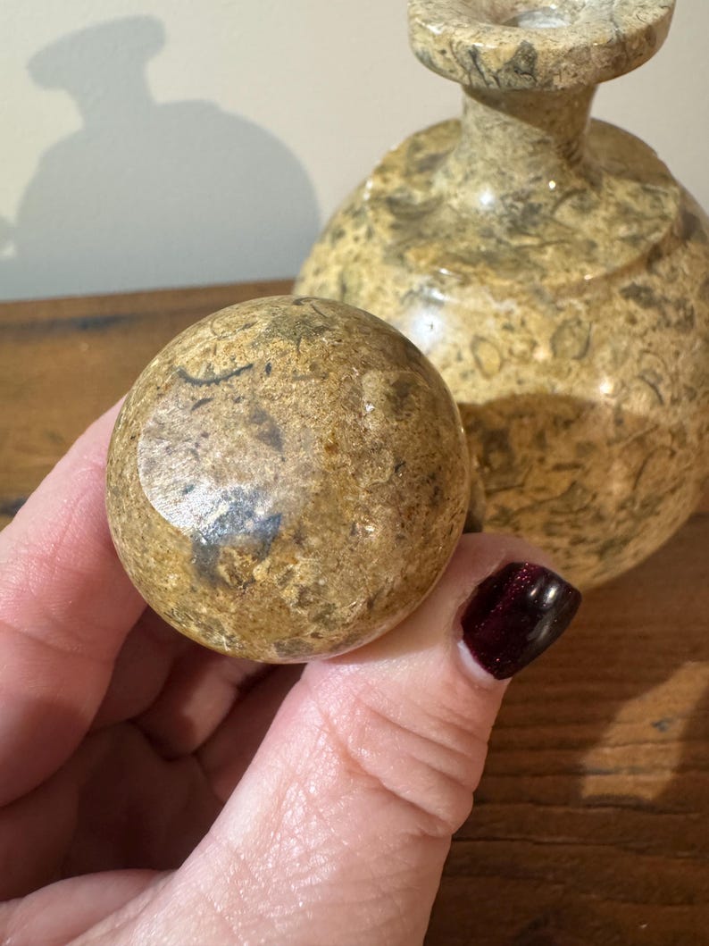 K&ouml;nnte beinhalten: Nahaufnahme einer runden, gesprenkelten Steinkugel, die in einer Hand gehalten wird, mit einer passenden dekorativen Vase im Hintergrund. Die Kugel und die Vase haben ein beige-braunes Farbschema mit poliertem Finish. Der Hintergrund ist unscharf.
