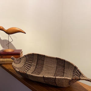May include: A woven basket with a leaf-like shape, featuring alternating bands of light and dark brown. The basket is displayed on a wooden shelf, alongside a wooden bird sculpture and a stack of vintage books.