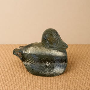 May include: A glass duck figurine with a textured surface. The duck is primarily a dark green color with white and black accents. The figurine is sitting on a tan surface.