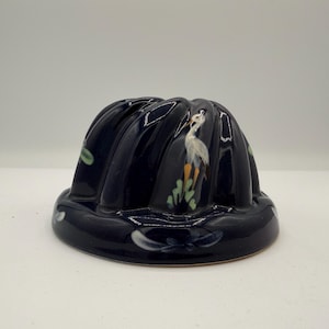 Pode incluir: Um molde canelado de cerâmica azul-marinho escuro com uma cegonha branca pintada à mão com pernas cor de laranja entre folhagem verde. O utensílio de cozinha brilhante e arredondado tem uma base larga e estrias verticais com um motivo.