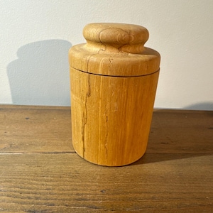 Peut inclure: Un récipient cylindrique en bois avec un couvercle arrondi. Le récipient est fait de bois de couleur claire avec des motifs de grain verticaux. Le couvercle est de forme plus petite et arrondie. Le récipient repose sur une surface en bois.
