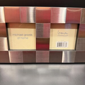 May include: A double picture frame with a brushed metal finish composed of square tiles. The left insert reads michael graves at home, and the right insert features a signature and a barcode with the text 3x5 inches printed above it.