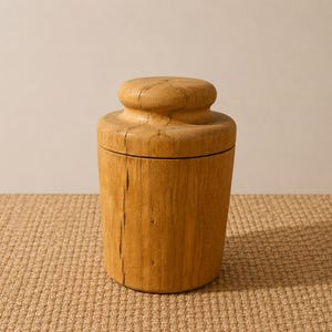 May include: A cylindrical wooden container with a rounded lid. The container is made of light brown wood with visible grain and natural imperfections. The lid is a similar color and shape, creating a cohesive design. The container sits on a textured, tan surface.