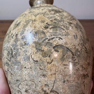 K&ouml;nnte beinhalten: Eine dekorative, steinartige Flasche mit rundem K&ouml;rper und kleinem, rundem Verschluss. Die Flasche hat ein gesprenkeltes Muster aus Beige, Braun und Grau mit fossilartigen Markierungen. Die Oberfl&auml;che ist glatt und poliert.