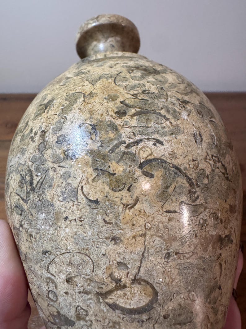 K&ouml;nnte beinhalten: Eine dekorative, steinartige Flasche mit rundem K&ouml;rper und kleinem, rundem Verschluss. Die Flasche hat ein gesprenkeltes Muster aus Beige, Braun und Grau mit fossilartigen Markierungen. Die Oberfl&auml;che ist glatt und poliert.