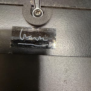 May include: A black textured surface featuring a silver-colored metal screw fastener at the top center. Below the fastener is a rectangular black label with a white handwritten signature that appears to read "Gann" with a horizontal line underneath.