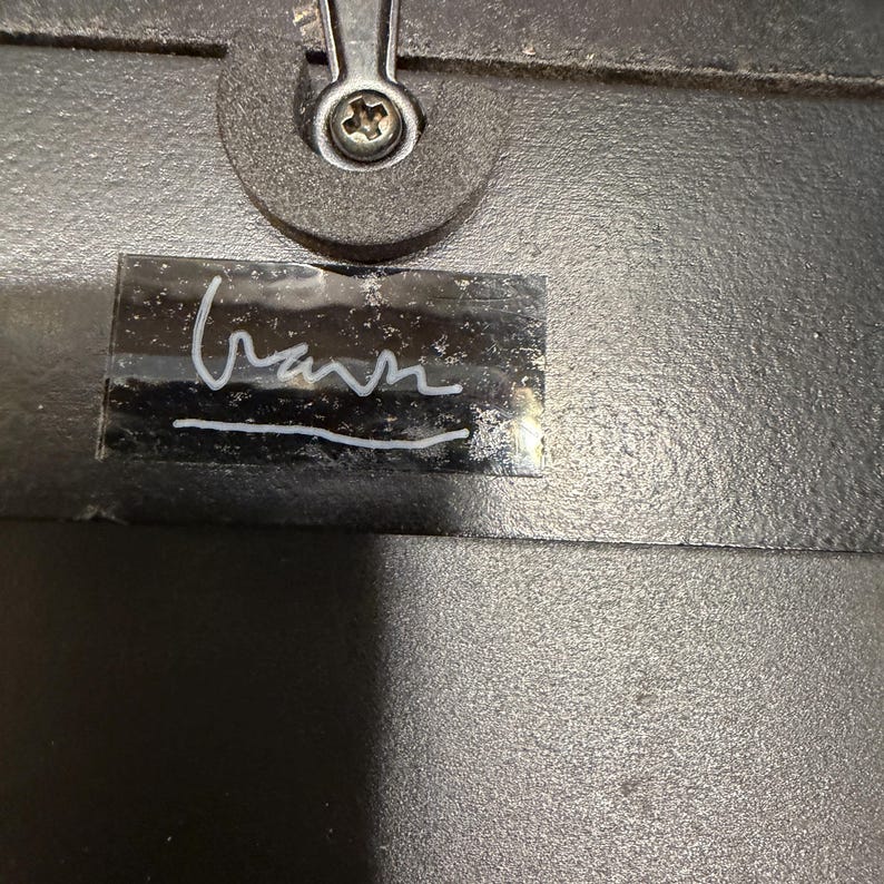 May include: A black textured surface featuring a silver-colored metal screw fastener at the top center. Below the fastener is a rectangular black label with a white handwritten signature that appears to read "Gann" with a horizontal line underneath.