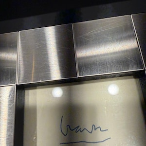 May include: A square picture frame featuring a border composed of brushed silver-toned metal tiles. The frame surrounds a light-colored insert that displays a handwritten signature in blue ink, positioned centrally within the frame's inner opening.
