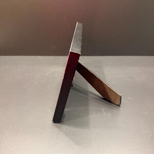 May include: A rectangular picture frame viewed from the side, featuring a thin black outer edge and a dark red inner border. The frame is supported by a brown, textured, hinged easel stand that extends outward to hold the frame in an upright position.