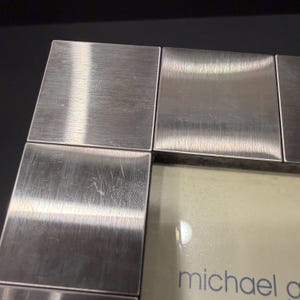 May include: A square-edged picture frame featuring a brushed silver-tone metal finish with a grid-like border design. The frame contains a light-colored insert with the partial text michael g printed in a simple, lowercase blue font at the bottom.