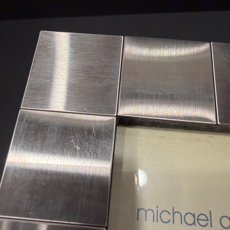 May include: A square-edged picture frame featuring a brushed silver-tone metal finish with a grid-like border design. The frame contains a light-colored insert with the partial text michael g printed in a simple, lowercase blue font at the bottom.