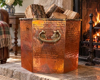 May include: A hexagonal copper log bucket with a hammered finish and two brass swing handles. The container features a riveted top rim and is filled with several pieces of split firewood, showcasing a metallic sheen and warm tones.