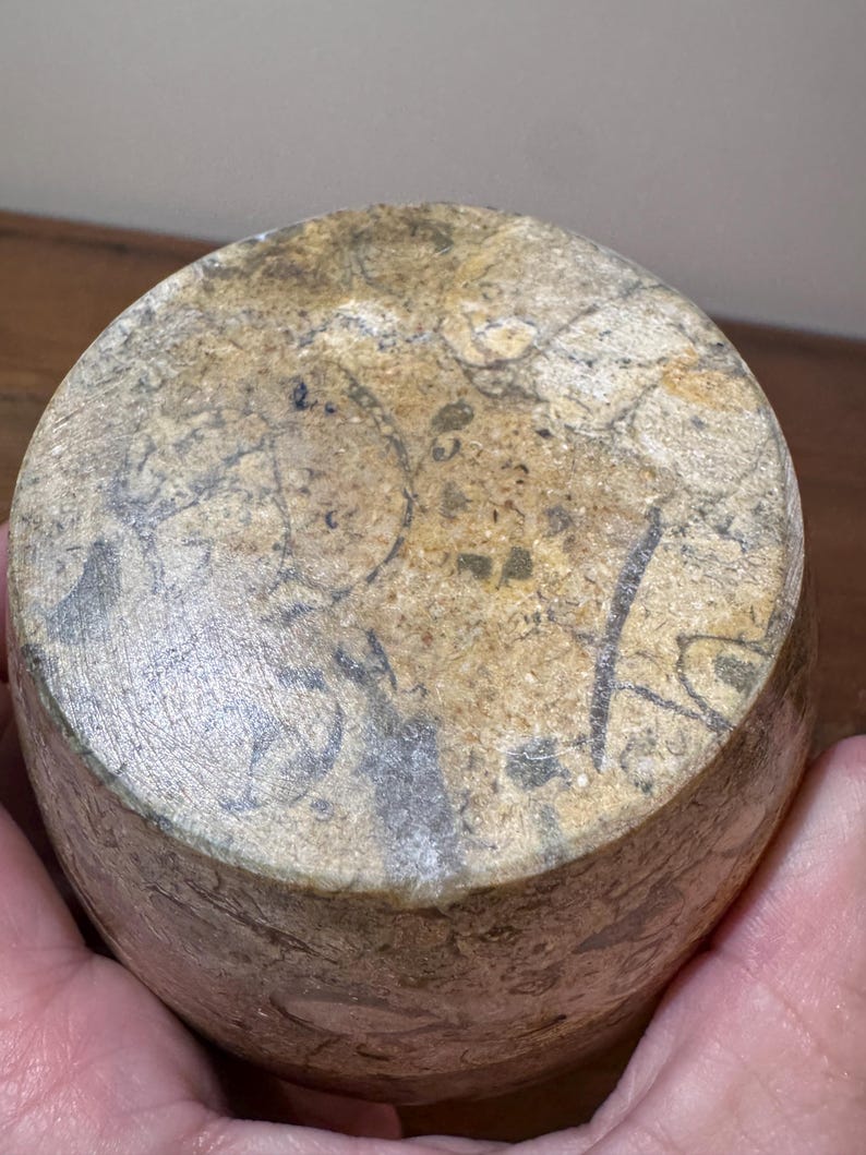 K&ouml;nnte beinhalten: Ein zylindrisches Steinobjekt mit einem gesprenkelten Muster aus Beige, Grau und Braun. Der Stein hat eine raue, strukturierte Oberfl&auml;che mit dunklen Markierungen. Das Objekt wird in einer Hand gehalten.