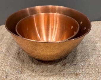 May include: Two nested copper bowls with a polished metallic finish. The bowls feature a rounded shape and visible rivets on the side of the larger bowl, showcasing the warm, reddish-brown tone of the copper material in a simple, functional design.