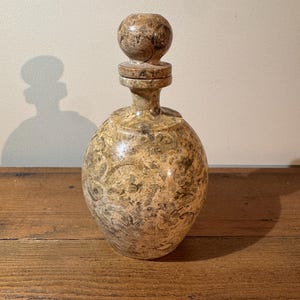 K&ouml;nnte beinhalten: Eine dekorative, runde Flasche mit passendem Verschluss. Die Flasche und der Verschluss bestehen aus einem gesprenkelten, hellbraunen Material mit braunen und beigen Mustern. Die Flasche steht auf einer Holzoberfl&auml;che.