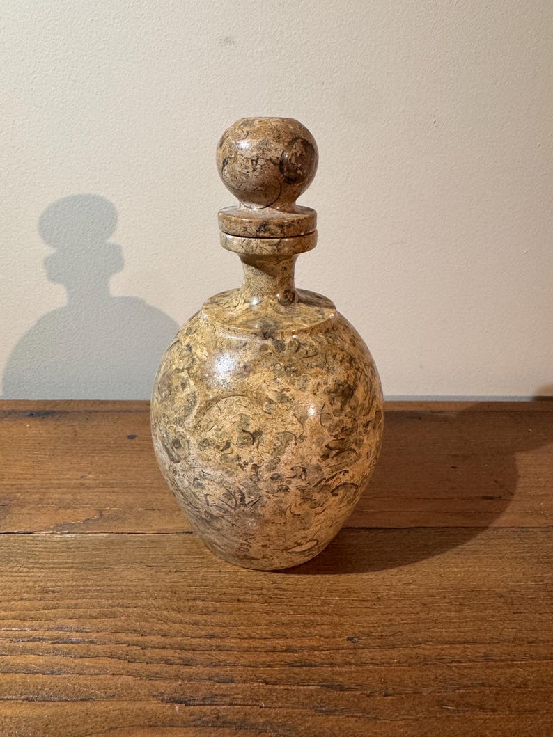 K&ouml;nnte beinhalten: Eine dekorative, runde Flasche mit passendem Verschluss. Die Flasche und der Verschluss bestehen aus einem gesprenkelten, hellbraunen Material mit braunen und beigen Mustern. Die Flasche steht auf einer Holzoberfl&auml;che.