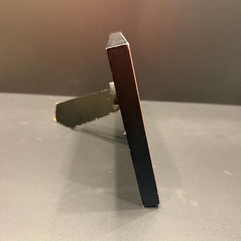 May include: A side view of a rectangular black picture frame featuring a thin profile and a metallic easel stand attached to the back. The frame is positioned at an angle, showing the dark edge and the mechanical hinge of the support structure.