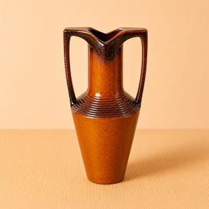 May include: A brown ceramic vase with a distinctive form. It features a broad, angular top with two handles and a tapered body. The surface has a glossy finish, transitioning from dark brown at the top to a lighter brown at the base. The vase has a ribbed texture around the middle.