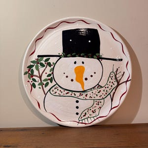 May include: A white ceramic plate featuring a hand-painted snowman design. The snowman has a black top hat, an orange carrot nose, and a dotted scarf. The plate's rim has a red wavy border and green holly accents.