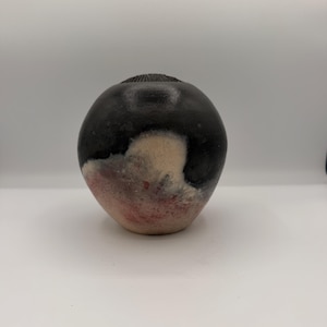 May include: A spherical ceramic vase with a smooth, matte black upper section and a textured, porous lower section featuring blended gradients of cream, white, and soft pink tones. The top opening is small and circular with a dark, ridged rim.