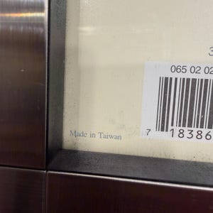 May include: A close-up view of a dark metallic frame edge bordering a cream-colored surface. A white rectangular sticker with a black barcode and the numbers 065 02 02 is attached to the surface. The text Made in Taiwan is printed in black.
