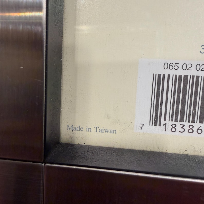 May include: A close-up view of a dark metallic frame edge bordering a cream-colored surface. A white rectangular sticker with a black barcode and the numbers 065 02 02 is attached to the surface. The text Made in Taiwan is printed in black.