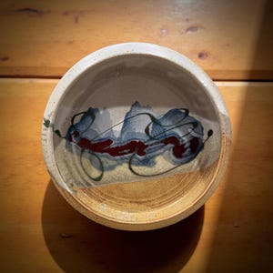 May include: A handmade ceramic bowl with a cream-colored rim and a tan exterior. The interior features a blue and red abstract design with black squiggles. The bowl is set on a wooden surface.