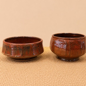 May include: Two handmade ceramic bowls with a rustic, textured appearance. The bowls are a deep reddish-brown color with darker accents, and have a faceted design. They sit on a textured, tan surface.