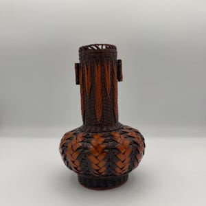 May include: A dark brown and reddish-brown woven bamboo vase featuring a wide, rounded base that tapers into a cylindrical neck. The neck is decorated with vertical leaf-like patterns, and two small, cylindrical bamboo handles are attached to the sides.