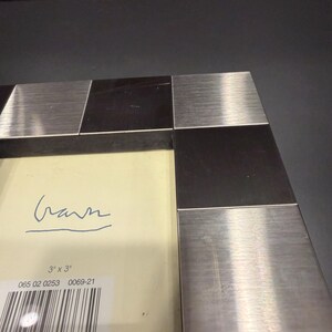 May include: A square picture frame featuring an alternating pattern of brushed silver-tone metal and dark brown wood blocks. The cream-colored insert displays the text 3" x 3", a barcode, and a handwritten signature in blue ink.