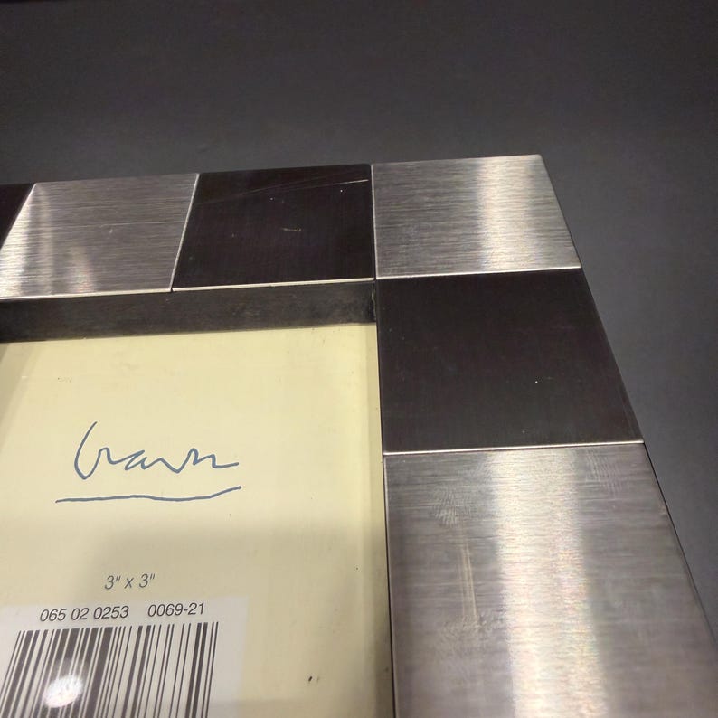 May include: A square picture frame featuring an alternating pattern of brushed silver-tone metal and dark brown wood blocks. The cream-colored insert displays the text 3" x 3", a barcode, and a handwritten signature in blue ink.