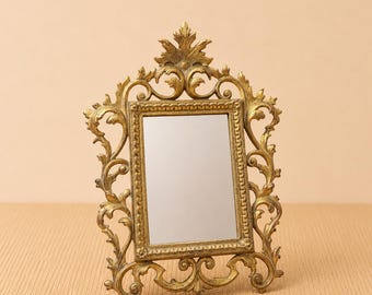 May include: A small, ornate gold-colored mirror with an elaborate, scrolling frame. The rectangular mirror is set within a detailed, vintage-style frame with a decorative top and base. The mirror is designed to stand upright on a flat surface.