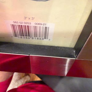 May include: A rectangular picture frame with a metallic silver border and a black inner edge. A white label is attached to the cream-colored backing, displaying the text 3" x 3", the numbers 065 02 0253 0069-21, and a barcode with the numbers 7 18386 81868 2.