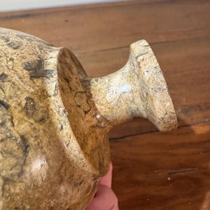 K&ouml;nnte beinhalten: Eine beige und braune dekorative Vase mit strukturierter Oberfl&auml;che. Die Vase hat einen abgerundeten K&ouml;rper und einen ausgestellten Rand. Die Vase besteht aus einem steinartigen Material.