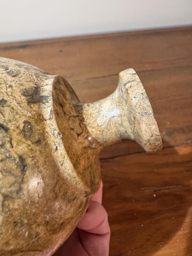 K&ouml;nnte beinhalten: Eine beige und braune dekorative Vase mit strukturierter Oberfl&auml;che. Die Vase hat einen abgerundeten K&ouml;rper und einen ausgestellten Rand. Die Vase besteht aus einem steinartigen Material.